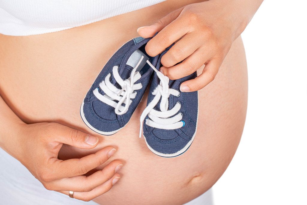 Pregnant woman holds small blue shoes on the her abdomen. Concept of happy pregnancy and expectation