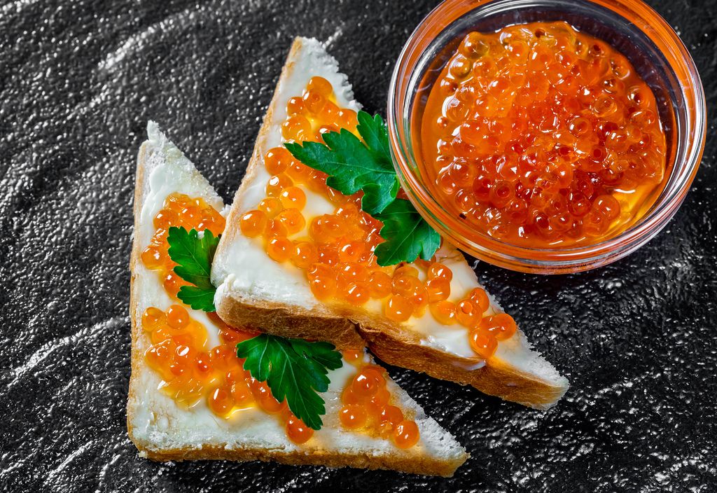Red caviar in a glass bowl and on sandwiches on a black stone background