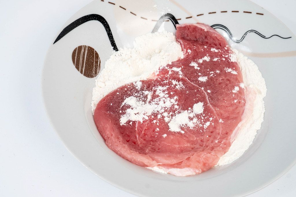 Rohes Schweinekotelett auf einem Teller mit Mehl vor weißem Hintergrund