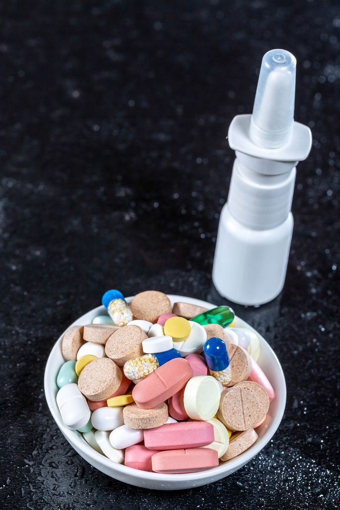Schale mit farbigen Pillen und Tabletten neben Nasenspray bei Erkältung vor schwarzem Hintergrund