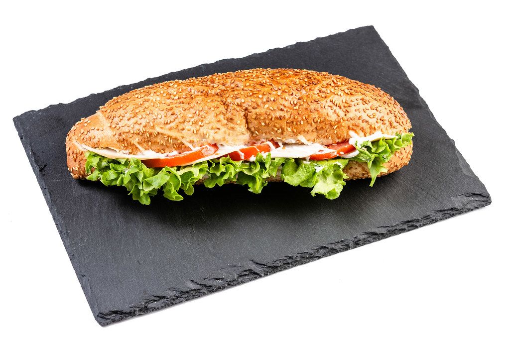 Sesambrötchen-Sandwich belegt mit Salatblätter, Schinken, Tomaten und weißer Sauce auf schwarzer Schieferplatte