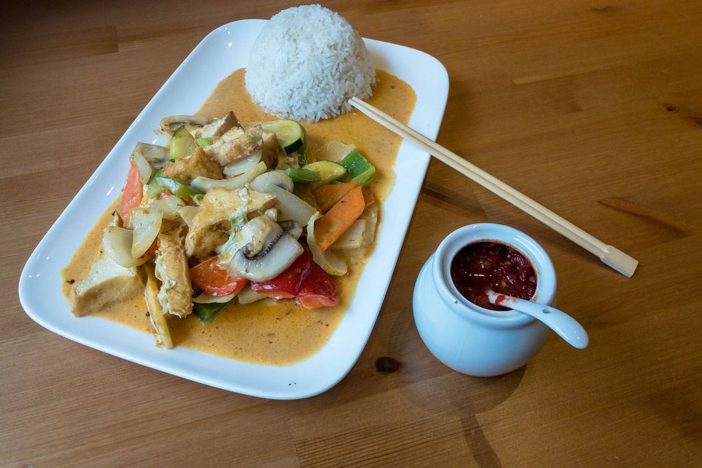 Thai Food - Rotes Vegan Curry mit Tofu, Pilzen, Zucchini, Paprika und Karotten mit geformten Reis auf einem Teller mit Bambus-Essstäbchen und Chilipaste in einem Gefäß