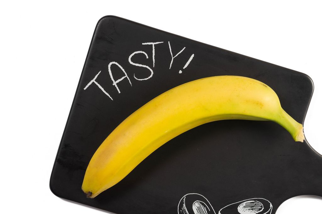 Ungeschälte, sattgelbe Banane auf schwarzem Küchenbrett mit Text TASTY vor weißem Hintergrund