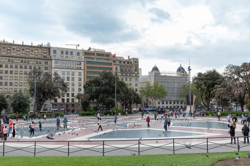 Verkehrsknotenpunkt Placa de Catalunya mit Tauben und Menschen auf sternförmigem Muster der rot-blauen Bodenfliesen in Barcelona, Spanien