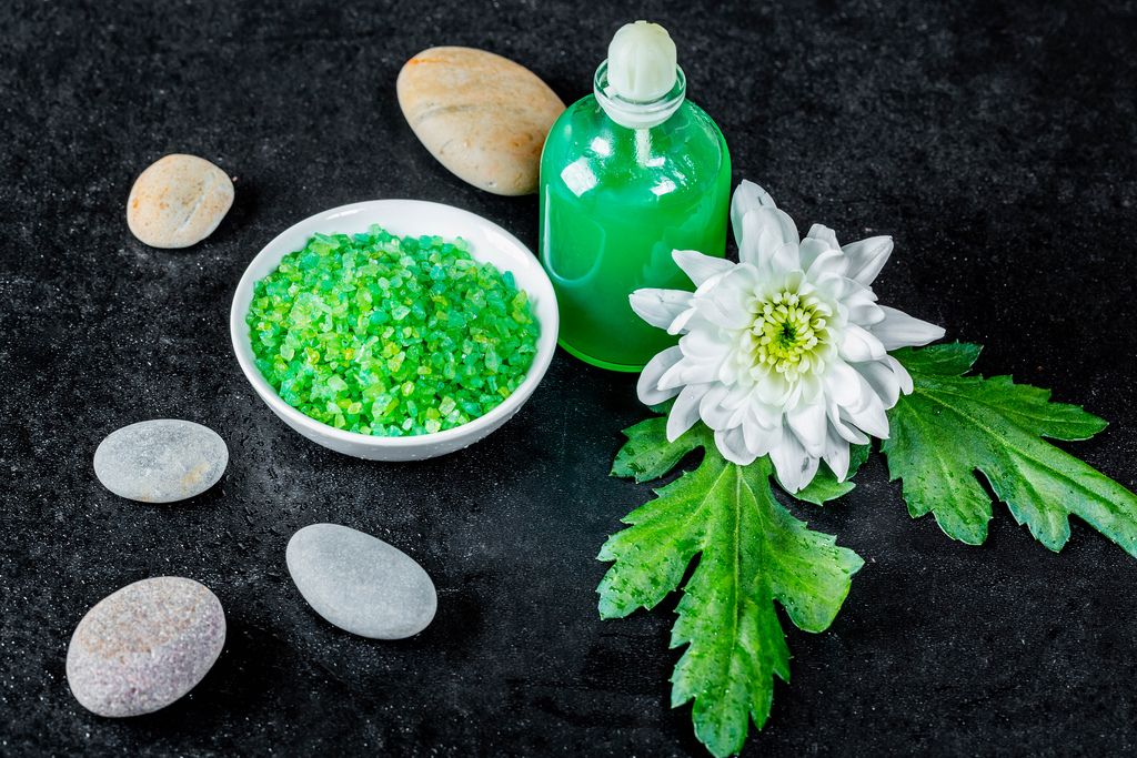 Wellnesszubehör wie Schale mit grünem Badesalz, grünes Gel und Steine mit Blume auf schwarz