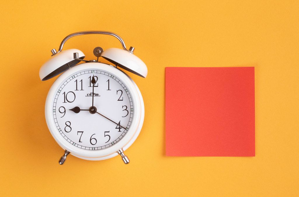 White alarm clock on yellow background with empty red sticky note