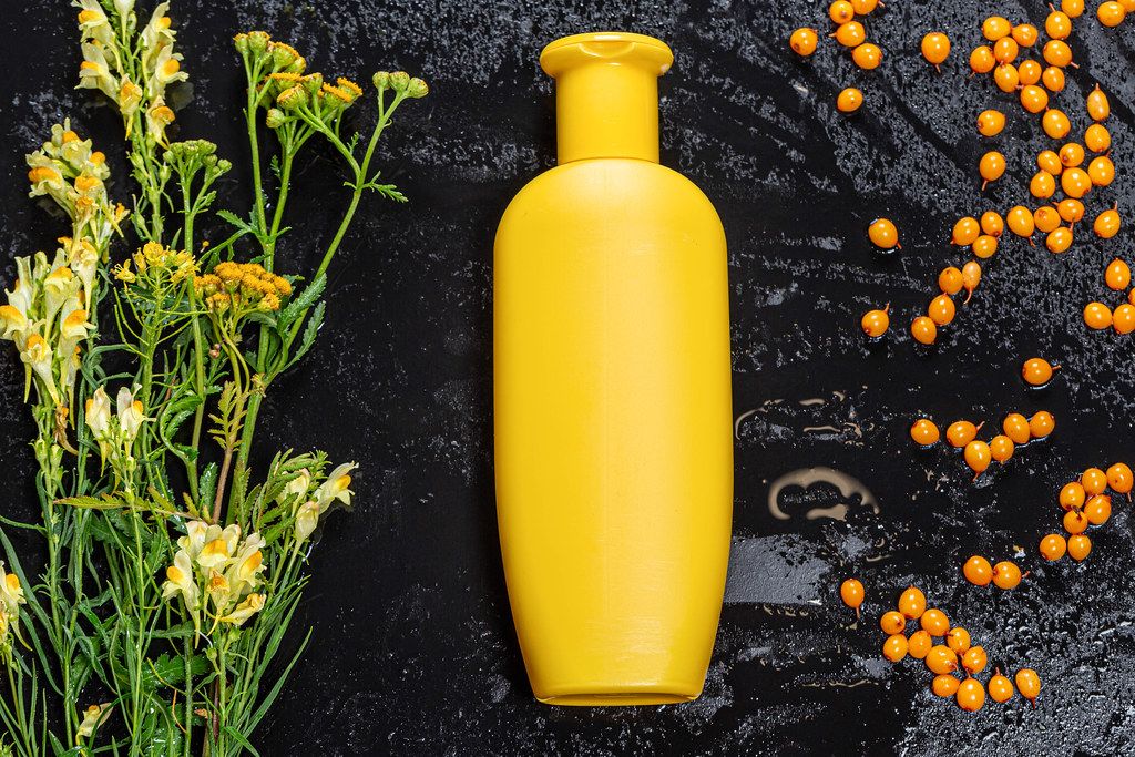 Yellow plastic bottle and sea buckthorn berries and wildflowers on black background. Natural care products