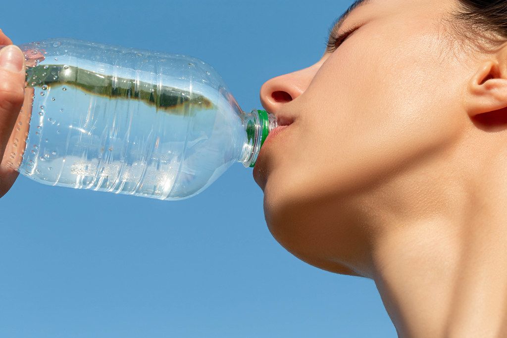 A girl drinks water from a plastic bottle on a blue sky background, close-up
