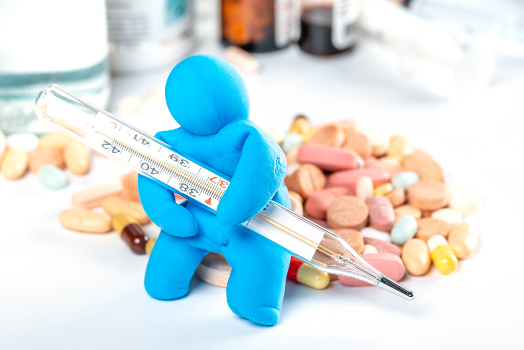 A man made of plasticine sits with a thermometer in his hands on a pile of pills