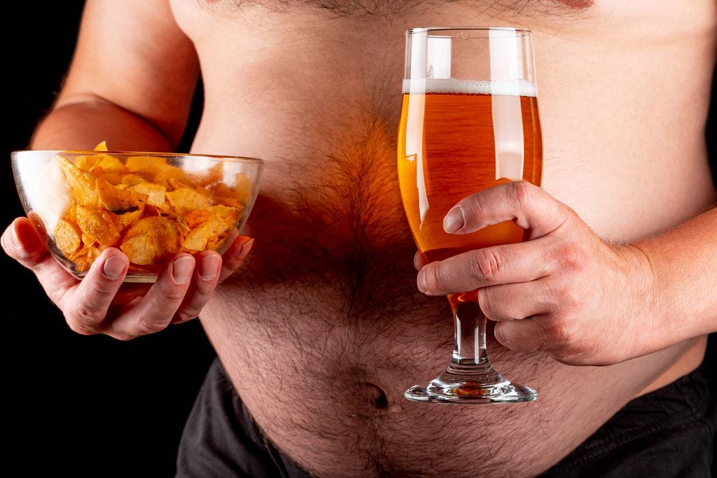 A man with a beer belly holds a glass of beer and chips