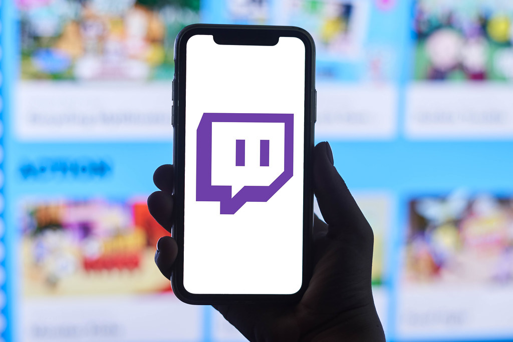 A person holds a smartphone with Twitch application logo over Game's Web Portal in the background