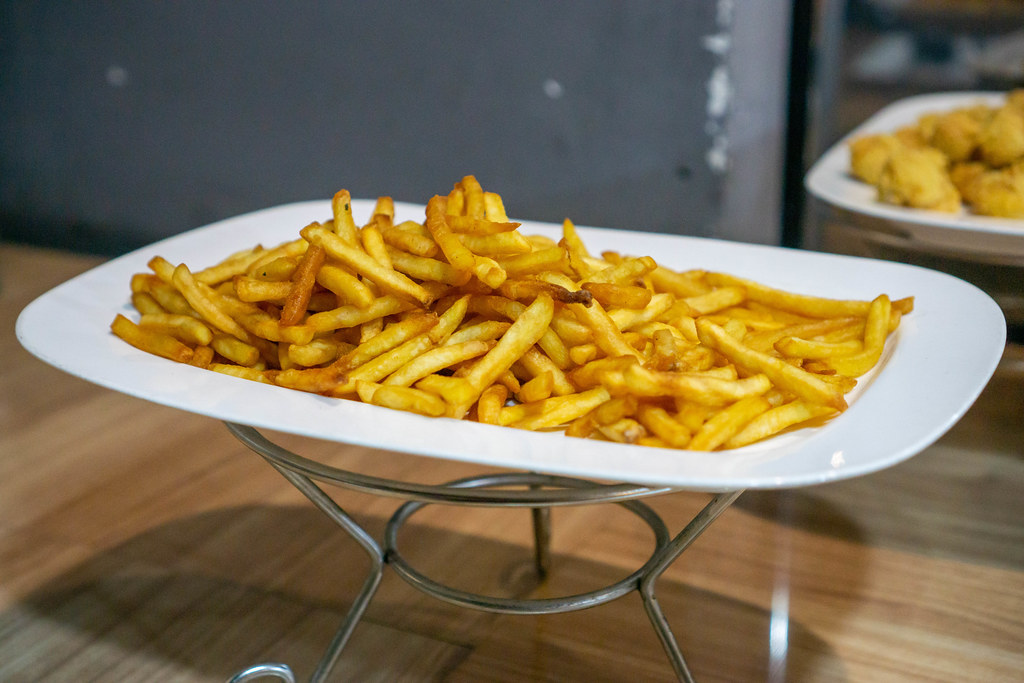 A Plate of Golden French Fries on a Metal Stand with Fried Chicken in the Background at a Catering Buffet
