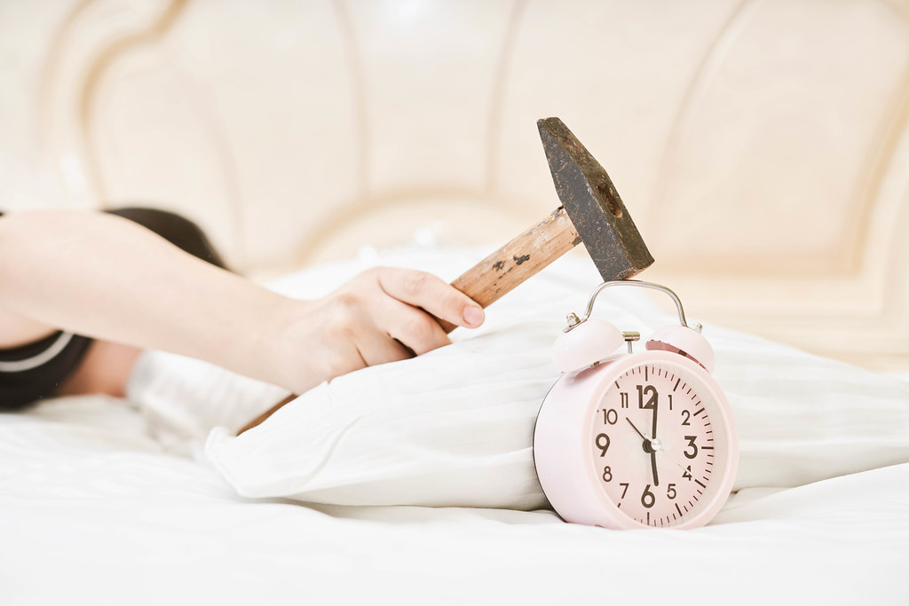 A sleeping woman in bed about to strike ringing alarm clock with a hammer