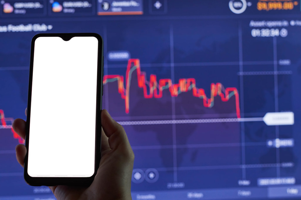 A trader holding a smartphone with an empty screen over the stock market live graphs