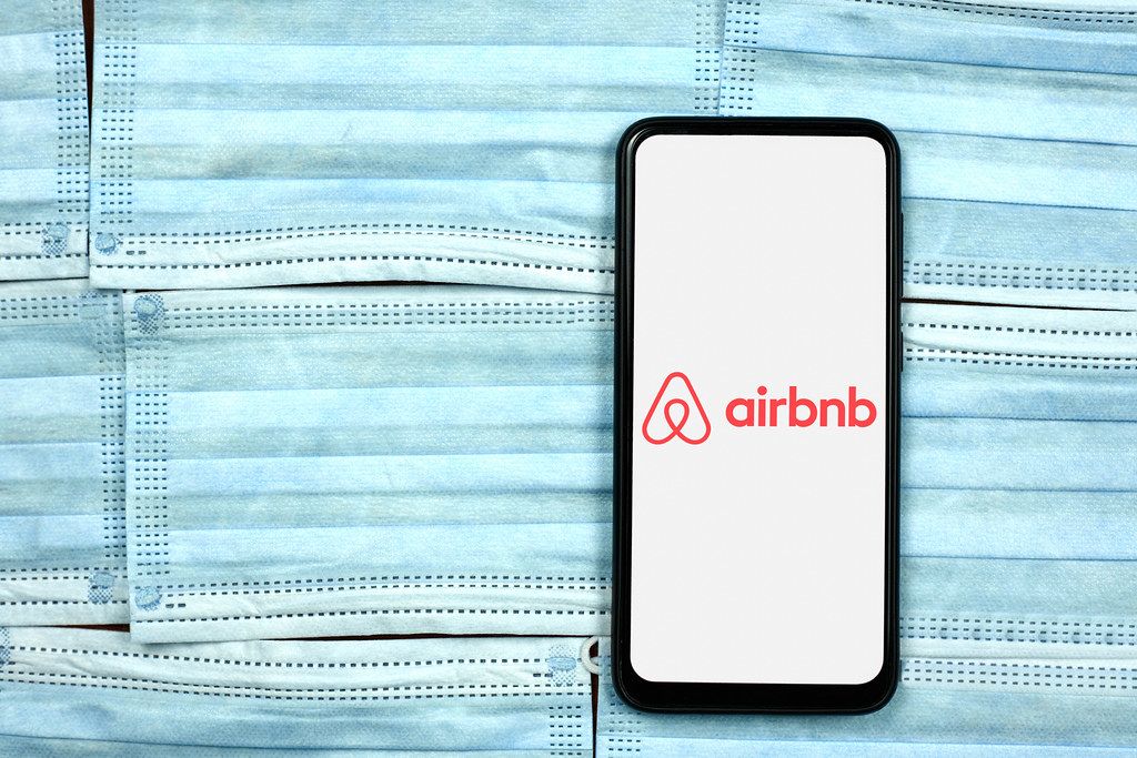 Airbnb company logo on smartphone screen over the face masks. Global company during coronavirus crisis