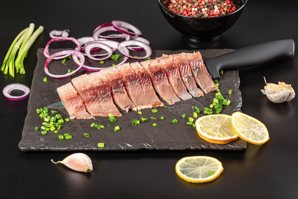 Appetizing pieces of pickled herring with red and purple onions, mix peas and sliced lemon on dark background