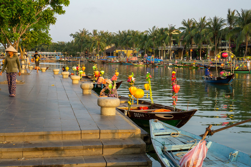 Bäume und Palmen entlang der Promenade am Flussufer vom Thu Bon Fluss mit vielen Ruderbooten dekoriert mit bunten Laternen für Touristen in Hoi An, Vietnam