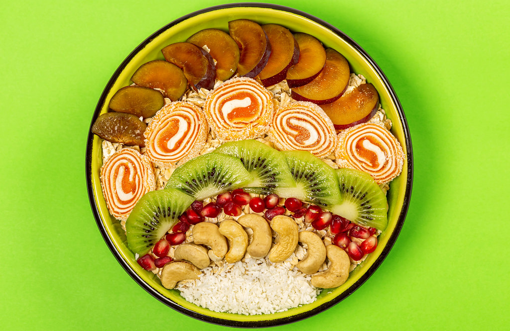 Bowl of oat flakes with plum pieces, turkish delight, kiwi, pomegranate, cashews and coconut on green, top view