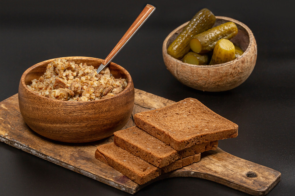 Buckwheat porridge in a wooden bowl on a dark background with pickles and black bread