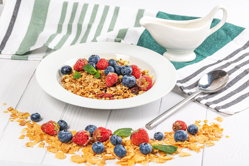 Cereal flakes, strawberries, blueberries and mint on a white wooden table with a spoon and a tea towel