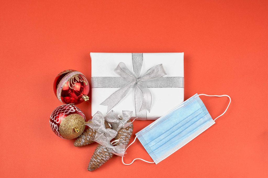 Christmas wrapped gift box, decorative items and face mask on red background