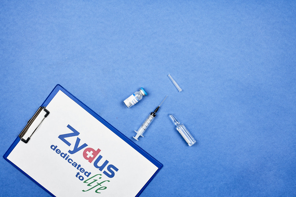 Clipboard with logo of Zydus Cadila, ampoule and syringe for Covid-19 vaccine on blue background