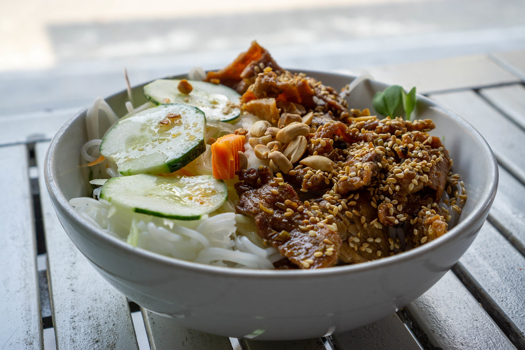 Close Up Food Photo of a Bowl of Vietnamese Bun Thit Nuong with Rice Vermicelli, Grilled Pork Meat, Sesame, Cucumber, Pickles and Peanuts on a White Wooden Table