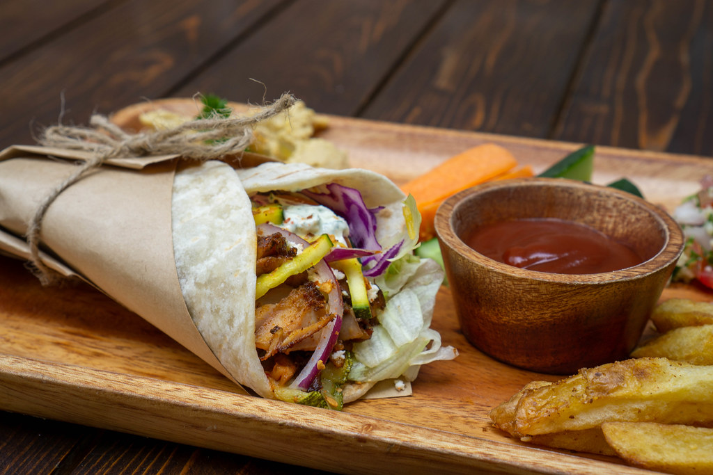 Close Up Food Photo of Doner Durum Kebab Roll with Chicken, Fresh Vegetables and Tztziki Sauce on a Wooden Lunch Plate with French Fries, Hummus and other Side Dishes