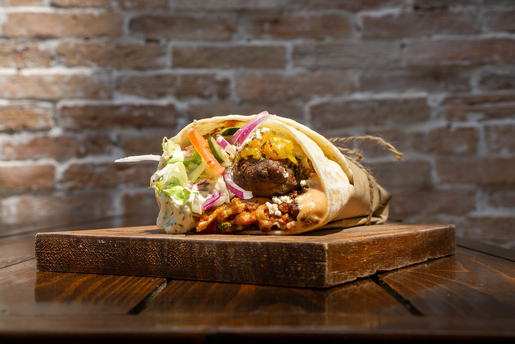 Close Up Food Photo of Doner Durum Kebab Roll with Lamb Meat, Raw Onions, Lettuce, Zucchini, Feta Crumbles, Tzatziki Sauce and Cheese on a Wooden Board with Brick Wall in the Background