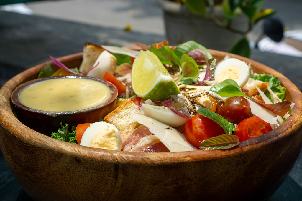 Close Up Food Photo of Healthy Mixed Salad Bowl with Chicken, Parmesan Slices, Boiled Qual Eggs, Basil, Cherry Tomatoes, Fried Bread Cubes, Lime and Honey Mustard Sauce in a Wooden Bowl