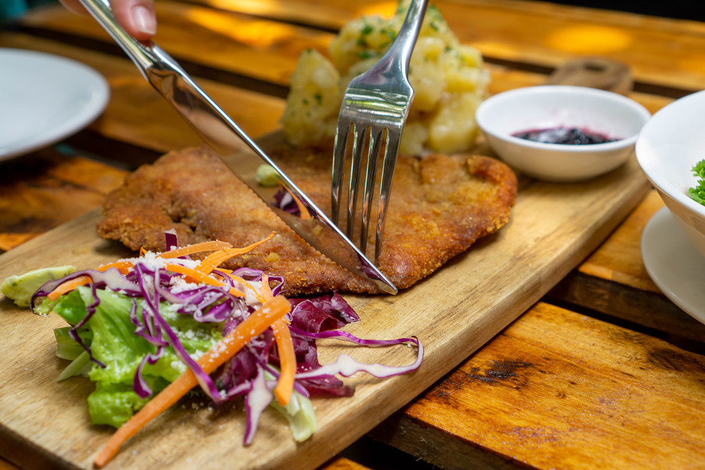 Close Up Food Photo of Knive and Fork being used to cut a Chicken Schnitzel on a Wooden Board with Homemade Potato Salad and Garden Salad in a German Restaurant