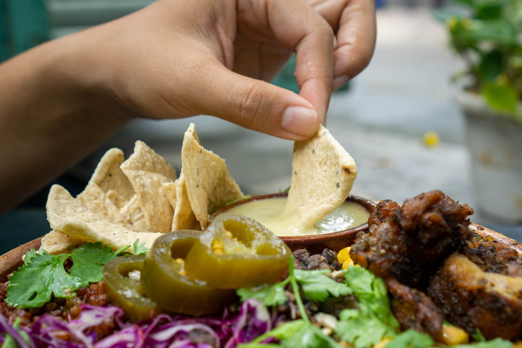 Close Up Food Photo of Person dipping a Tortilla Chip in Honey Mustard Sauce with a Mixed Salad Bowl with Grilled Chicken, Jalapenos, Cabbage and Herbs in the Foreground