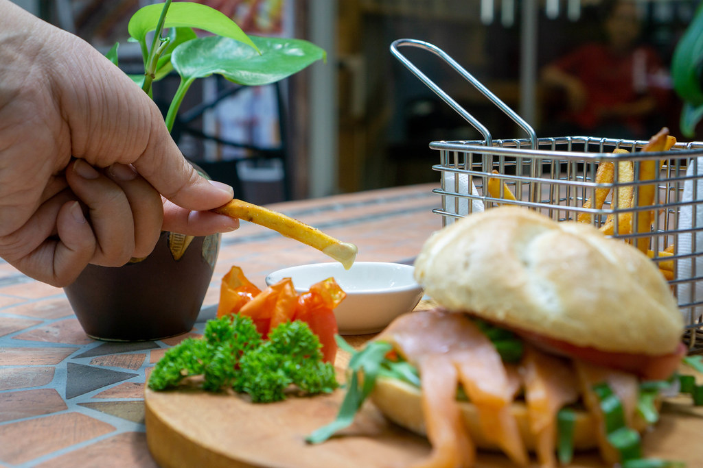 Close Up Food Photo of Person dipping French Fries in Honey Mustard Sauce with Smoked Salmon Sandwich on a Wooden Board in the Foreground