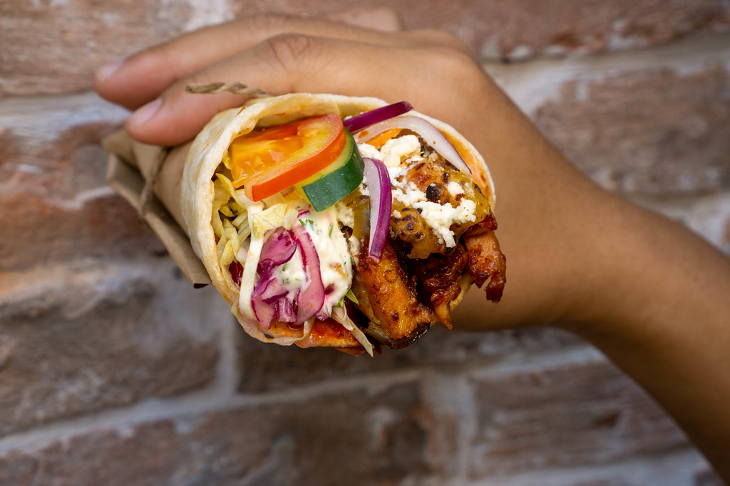Close Up Food Photo of Person holding a Doner Durum Wrap with Chicken, Tomato, Cucumber, Red Cabbage, Lettuce, Onions and Feta Crumble in front of a Brick Wall