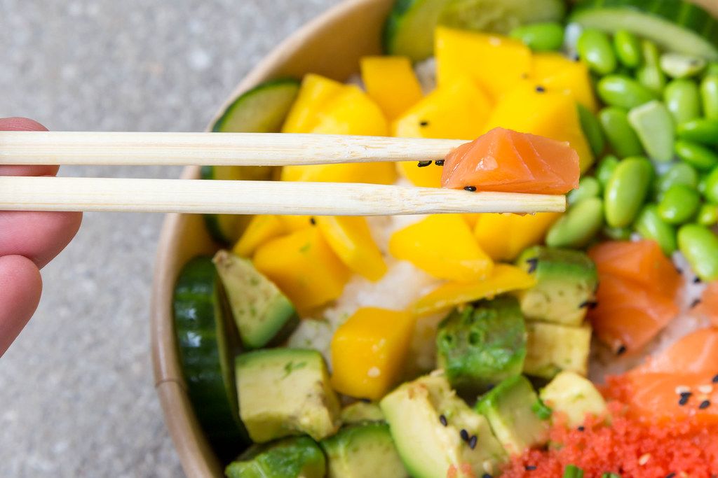 Close-up: hand with chopsticks and piece of salmon from a poké bowl with mango, avocado, soy beans