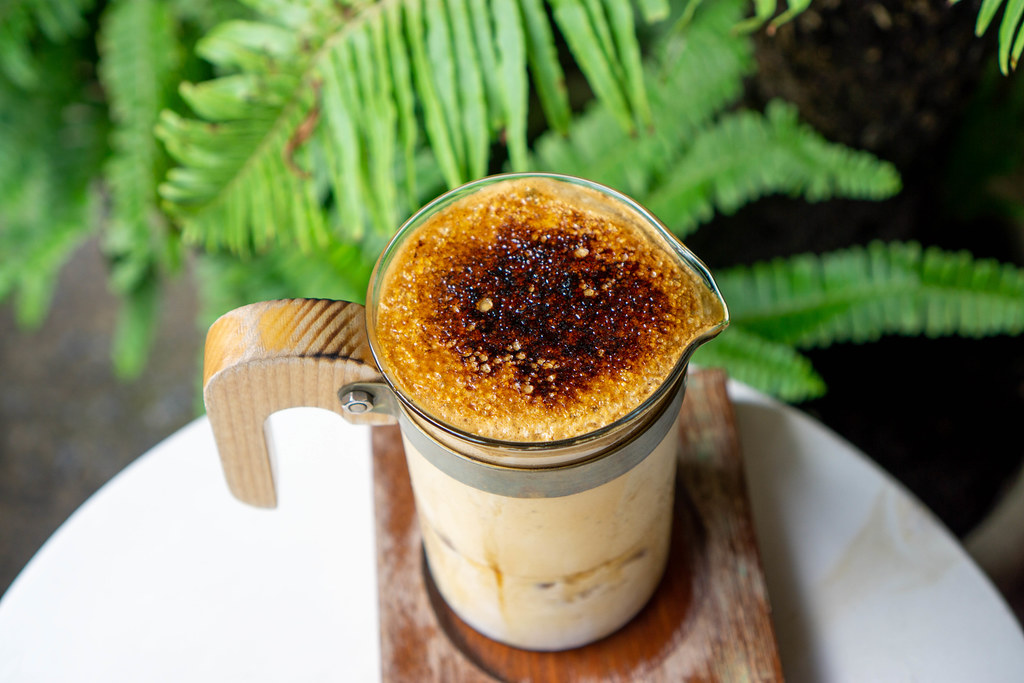 Close Up Photo of Burnt Caramelized Sugar on Top of a Dalgona Coffee in a Designer Coffee Glas with Wooden Handle with Plants in the Background