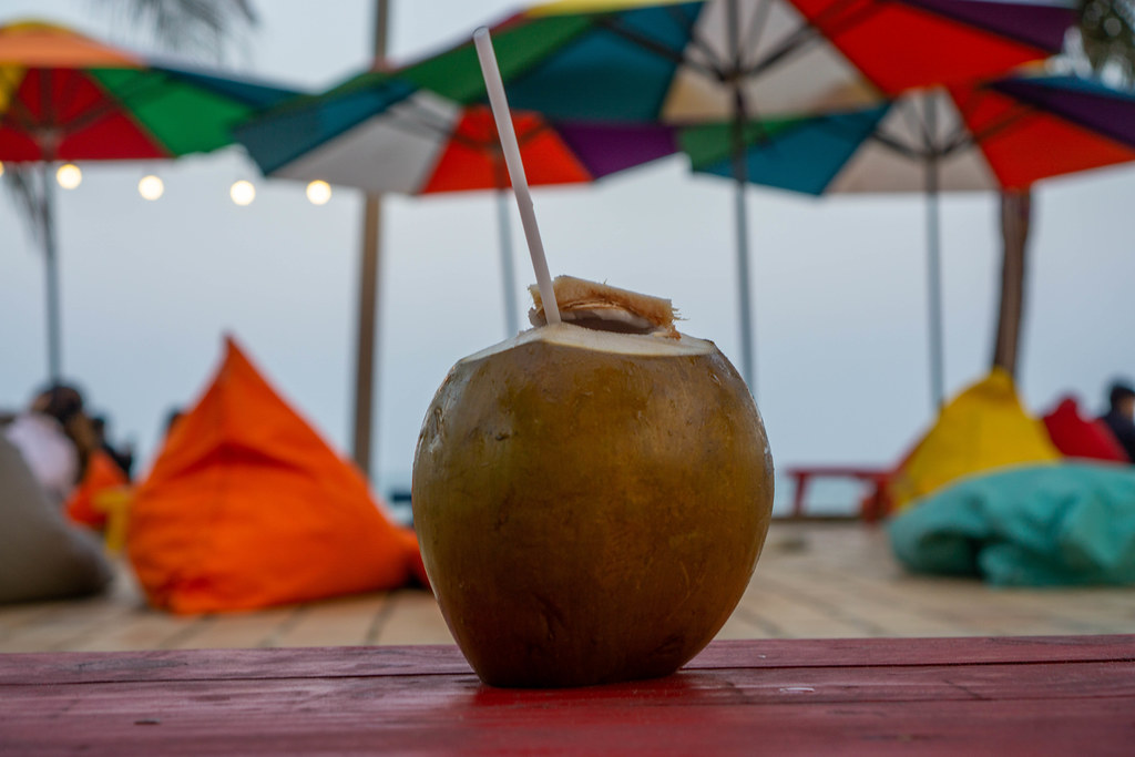 Close Up Photo of Fresh Coconut with Drinking Straw on a Wooden Table with Sun Umbrellas and Bean Bags of a Beach Bar in the Background
