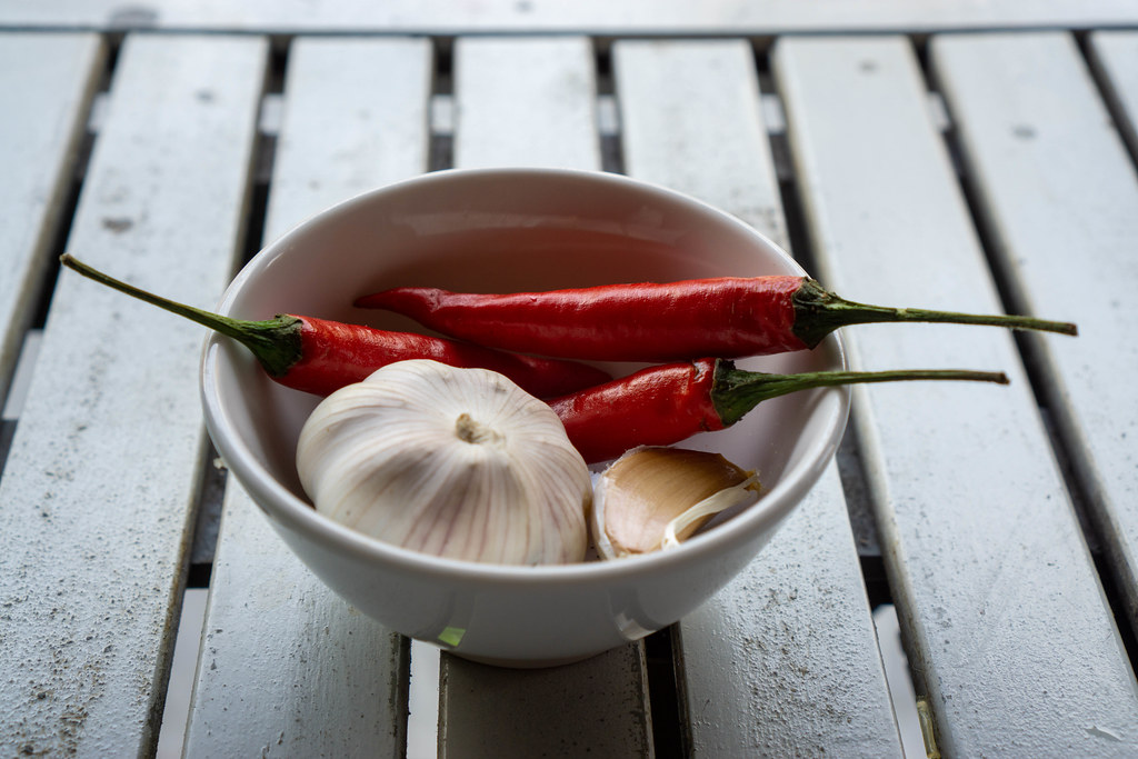 Close Up Photo of Fresh Thai Red Chilis in a Small Ceramic Bowl with Garlic Cloves and a Garlic Bulb on a White Wooden Table