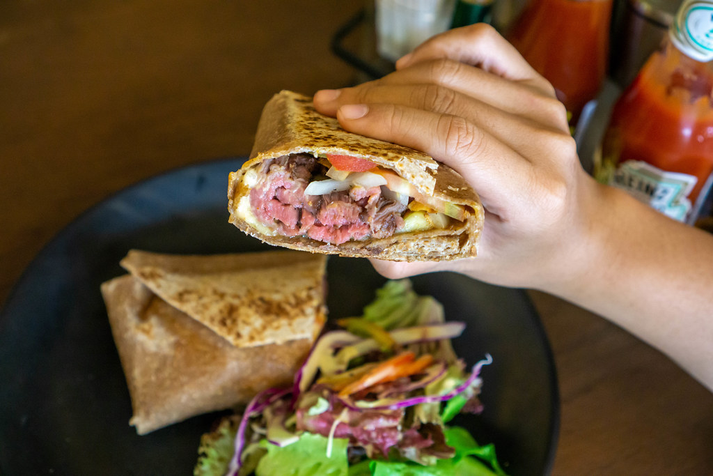Close Up Photo of Person holding a Tortilla Wrap with Roast Beef, Melted Gouda Cheese and Vegetables with Plate and Sauces in the Background