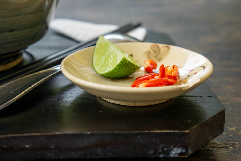 Close Up Photo of Piece of Lime and Fresh Chopped Chili on a Small Ceramic Plate next to a Spoon and Chopsticks on a Wooden Tray