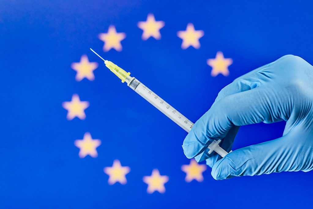Corona-Impfstoff: Hand mit Einmalhandschuhen hält eine Spritze vor der Flagge der Europäische Union