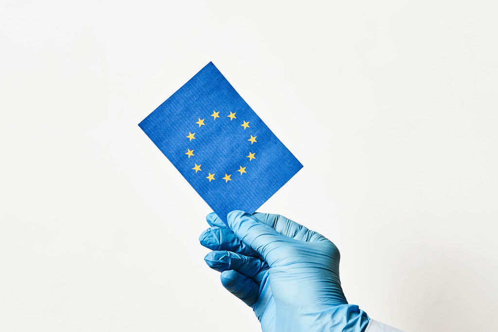 Corona und EU: Hand mit Einmalhandschuh hält eine europäische Flagge vor weißem Hintergrund