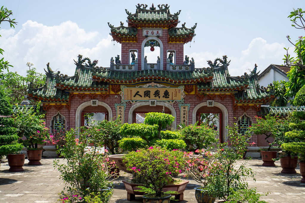 Courtyard with many different Plants and Trees in front of a Stone Entrance Gate with large Chinese Letters, Ornaments and Statues at the Phuc Kien Pagoda in Hoi An, Vietnam