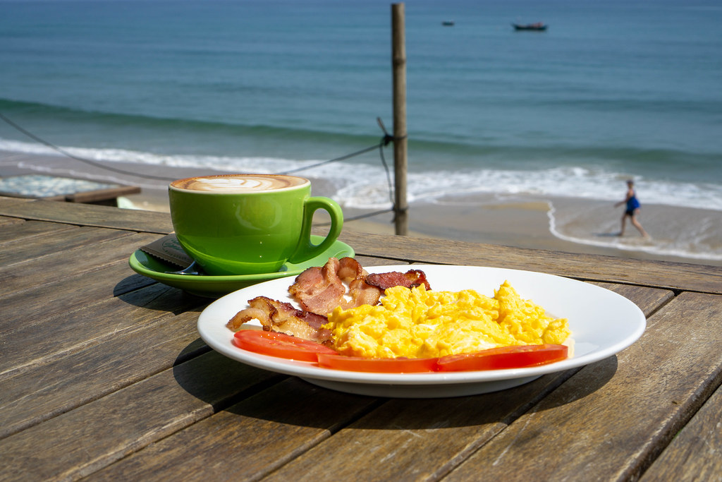 Cup of Cappuccino and Scrambled Eggs with Tomatoes and Bacon Strips for Breakfast in the Sun at An Bang Beach in Hoi An, Vietnam