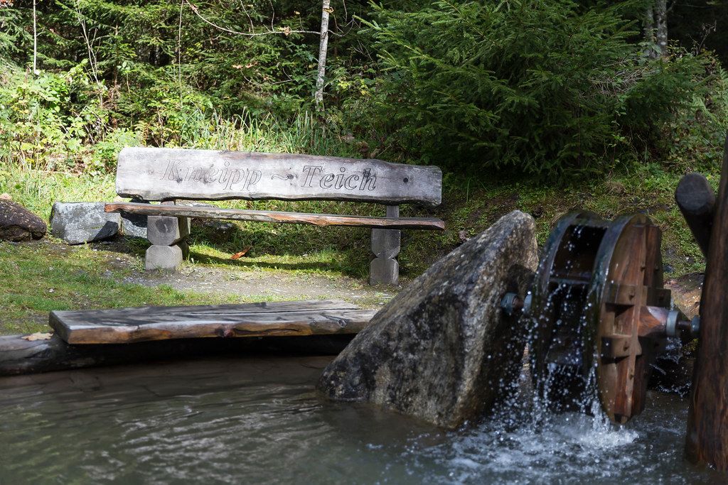 Eine Bank mit der Schrift "Kneipp - Teich" beim kleinen Teich mit Wasserlauf und Mühlrad in Alpbach