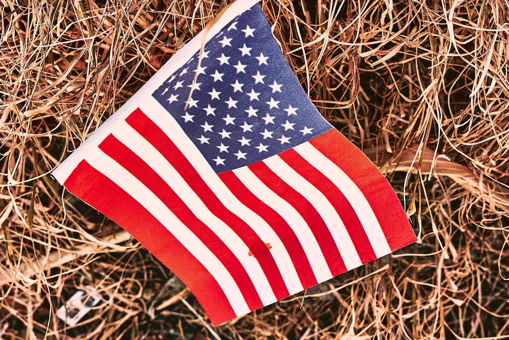 Eine US-Amerikanische Flagge mit einem Blutfleck liegt im Stroh. US-Politik im Chaos