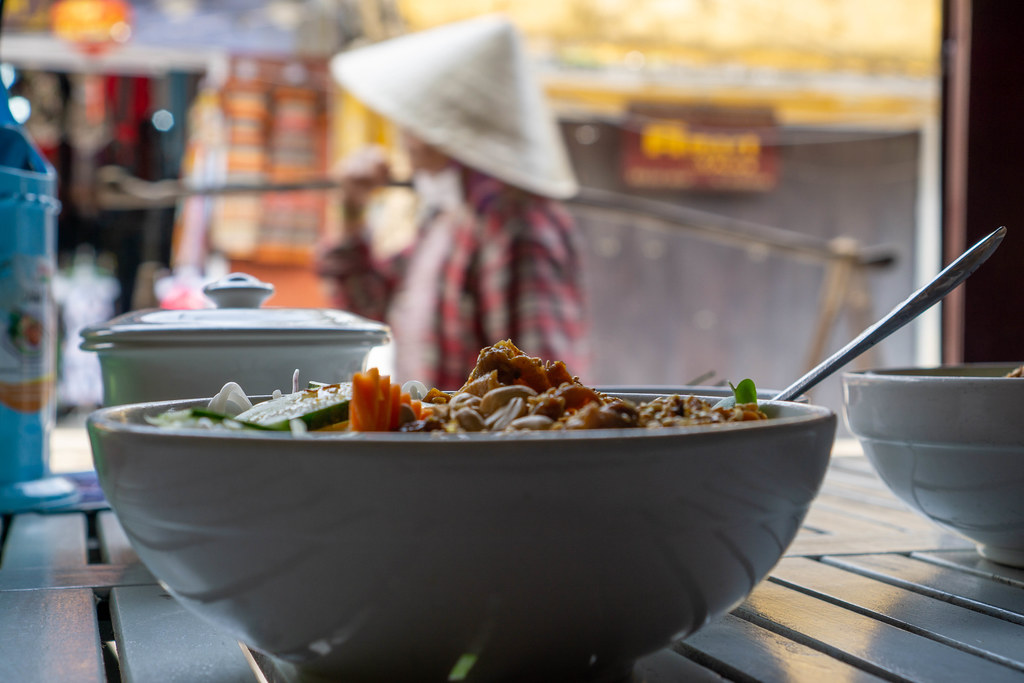 Food Photo of Bowl of Vietnamese Bun Thit Nuong with Vietnamese Woman with Conical Hat and Shoulder Pole in the Background in Hoi An, Vietnam