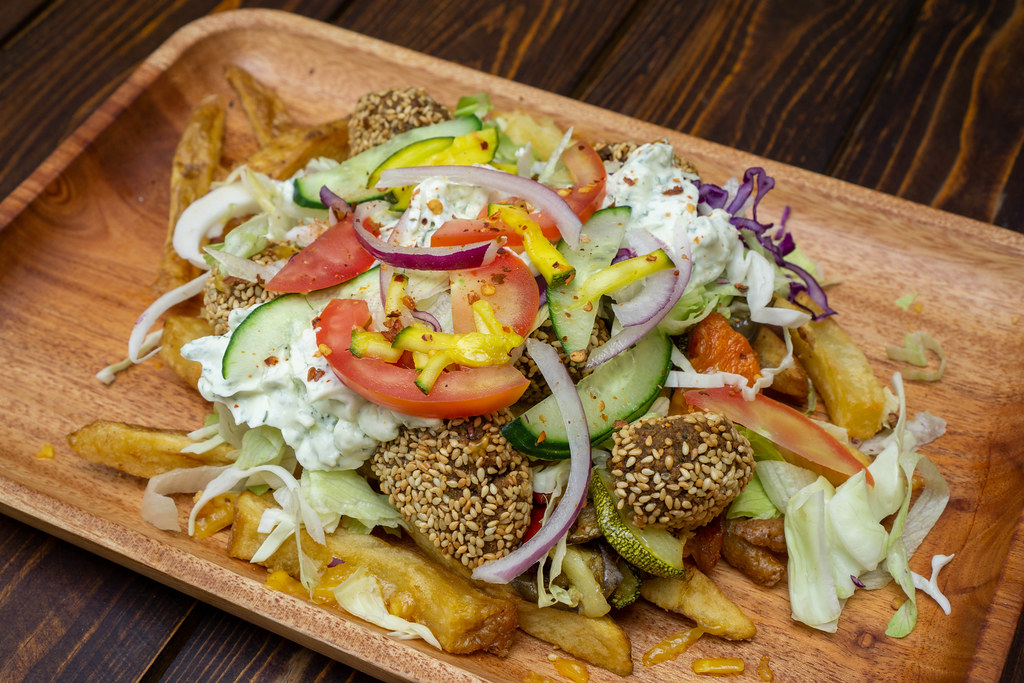 Food Photo of Kapsalon Salad with French Fries topped with Sesame Falafel, Lettuce, Onions, Zucchini, Cucumber and Tzatziki on a Wooden Tray at a Kebab Restaurant
