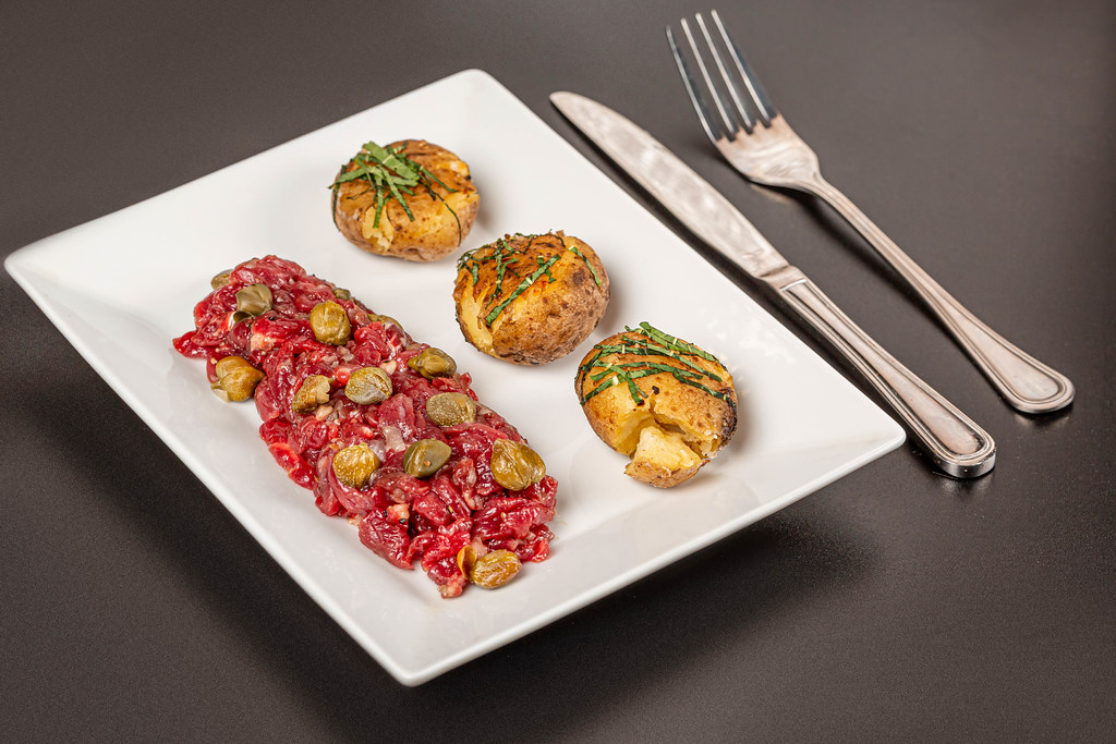 Fresh beef tartare with capers and potato on plate, dark background