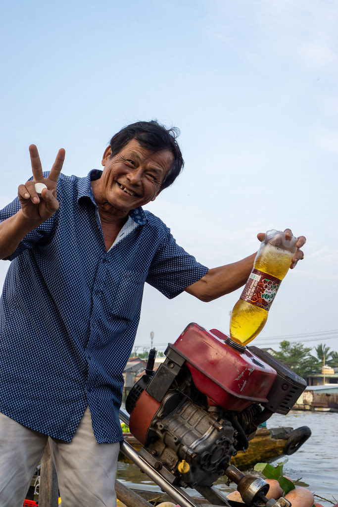 Freundlicher Mann tankt sein Motorboot mit Benzin aus einer Plastikflasche und lacht in die Kamera auf dem Cai Rang Schwimmenden Markt in Can Tho, Vietnam